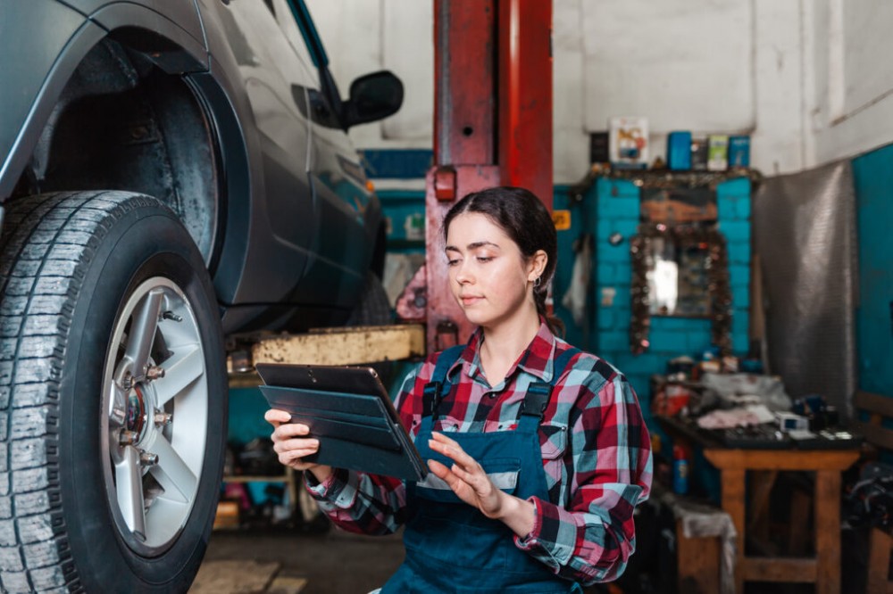 Portrait of a young female mechanic in uniform posing with a tablet in her hands. In the background is an auto repair shop. The car on the lift to the right.